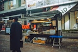 A small food stall located on a bustling city street, with a vendor standing inside arranging bottles and snacks. The surrounding area features modern shops and advertisements, indicating an urban setting. A person in a black coat is walking past, carrying a briefcase. The ground is sprinkled with fallen leaves, suggesting autumn.