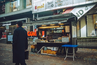 A small food stall located on a bustling city street, with a vendor standing inside arranging bottles and snacks. The surrounding area features modern shops and advertisements, indicating an urban setting. A person in a black coat is walking past, carrying a briefcase. The ground is sprinkled with fallen leaves, suggesting autumn.