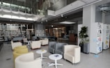 A spacious, modern library with high ceilings and ample natural light. The seating area consists of multiple armchairs and small tables arranged in a lounge-like manner. Shelves filled with books line the back wall, and there is a large bookshelf closer to the front. Potted plants add a touch of greenery. The overall interior design is sleek and minimalist with neutral tones.