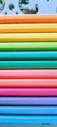 A neatly arranged stack of colorful paper or plastic sheets, each a different vibrant hue. The sheets are aligned horizontally, creating a rainbow effect from top to bottom. The top sheet has decorative cutout designs resembling puzzle pieces.