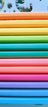 A neatly arranged stack of colorful paper or plastic sheets, each a different vibrant hue. The sheets are aligned horizontally, creating a rainbow effect from top to bottom. The top sheet has decorative cutout designs resembling puzzle pieces.