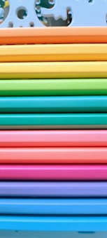 A neatly arranged stack of colorful paper or plastic sheets, each a different vibrant hue. The sheets are aligned horizontally, creating a rainbow effect from top to bottom. The top sheet has decorative cutout designs resembling puzzle pieces.