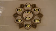 A decorative wall clock with a flower-like pattern, featuring multiple digital time displays and a central analog display showing a date. The frame has intricate designs with leaf shapes surrounding the displays, presenting a blend of modern digital and traditional decorative elements.