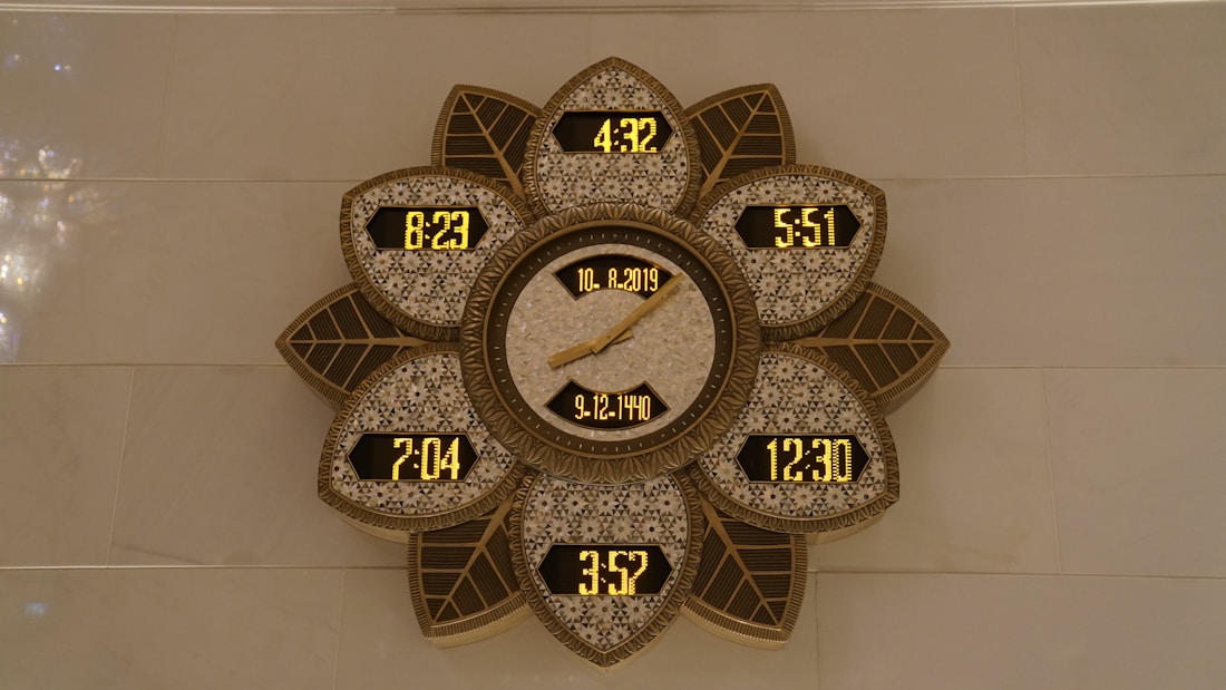 A decorative wall clock with a flower-like pattern, featuring multiple digital time displays and a central analog display showing a date. The frame has intricate designs with leaf shapes surrounding the displays, presenting a blend of modern digital and traditional decorative elements.