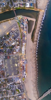 An aerial view of a beachfront amusement park with roller coasters and various attractions. The park is situated next to a sandy beach with waves gently lapping the shore. Residential areas and parking lots are visible alongside a body of water near the top of the image.