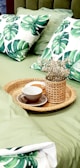 A cozy bedroom scene with a focus on a bed dressed in green-themed bedding. Two pillows with a large green leaf pattern rest on the green comforter. On the bed is a rattan tray holding a small, decorative vase filled with dried flowers and a cup with a saucer.