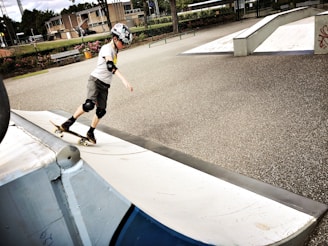 A young skateboarder wearing a helmet, elbow, and knee pads is practicing on a skate park ramp. The park features smooth concrete surfaces and other ramps in the background. There are some benches and greenery surrounding the area, and the skateboarder appears to be in motion, showcasing balance and focus.