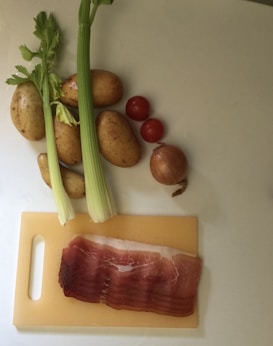 A collection of cooking ingredients including potatoes, celery stalks, cherry tomatoes, an onion, and slices of prosciutto are arranged on a light-colored surface. The prosciutto is placed on a small cutting board.