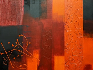 The image is an abstract painting featuring dominant vertical lines in varying shades of orange, red, and black. The painting has a mix of smooth and textured surfaces with splashes of orange paint scattered across the left side. The textures give depth to the different sections, while the interplay of dark and bright colors creates a vivid contrast.