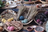 A collection of woven baskets containing various natural materials is arranged on a patterned cloth. The baskets hold yarn balls in different colors, dried herbs, seeds, and ears of dark seed corn. Additional elements include bowls with ground powders and scattered leaves in the background.