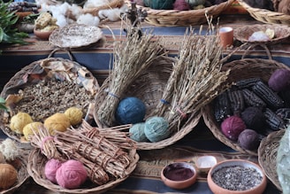 A collection of woven baskets containing various natural materials is arranged on a patterned cloth. The baskets hold yarn balls in different colors, dried herbs, seeds, and ears of dark seed corn. Additional elements include bowls with ground powders and scattered leaves in the background.
