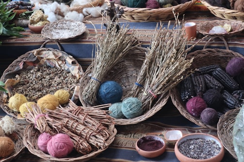 A collection of woven baskets containing various natural materials is arranged on a patterned cloth. The baskets hold yarn balls in different colors, dried herbs, seeds, and ears of dark seed corn. Additional elements include bowls with ground powders and scattered leaves in the background.