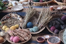 A collection of woven baskets containing various natural materials is arranged on a patterned cloth. The baskets hold yarn balls in different colors, dried herbs, seeds, and ears of dark seed corn. Additional elements include bowls with ground powders and scattered leaves in the background.