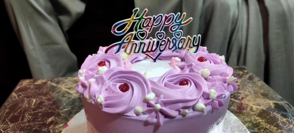 A round cake with purple frosting decorated with red cherries and white cream accents. A colorful 'Happy Anniversary' topper sits on the cake, which is placed on a marble-patterned surface with a neutral-colored background.