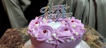 A round cake with purple frosting decorated with red cherries and white cream accents. A colorful 'Happy Anniversary' topper sits on the cake, which is placed on a marble-patterned surface with a neutral-colored background.