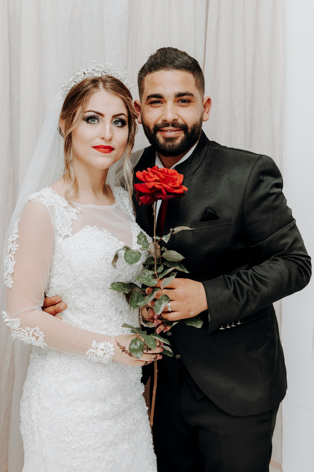 man in black suit jacket beside woman in white floral wedding dress holding red rose bouquet