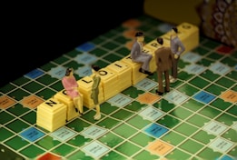 Miniature figurines are positioned on a Scrabble board, with yellow tile stacks spelling out 'NO L...ING'. The scene appears playful and imaginative, as the miniatures mimic real-life interactions in a board game setting.