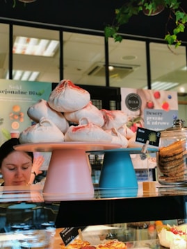 A bakery display featuring stacked meringues with a dusting of red powder on two cake stands, one pink and one blue. To the right, a jar filled with cookies is visible. The background shows a person with dark hair and an interior with posters and lights.