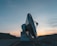 A large satellite dish is positioned on a concrete platform against a backdrop of a mountainous landscape. The sky is partly cloudy with warm hues from the setting sun.