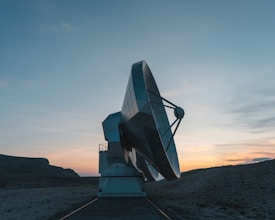 A large satellite dish is positioned on a concrete platform against a backdrop of a mountainous landscape. The sky is partly cloudy with warm hues from the setting sun.