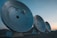 Three large satellite dishes are aligned in a row on a barren landscape. The sky above is clear with hints of sunset colors.
