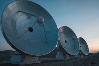 Three large satellite dishes are aligned in a row on a barren landscape. The sky above is clear with hints of sunset colors.