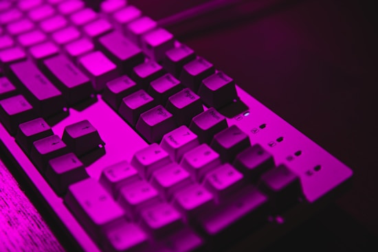 A keyboard illuminated by a vibrant pink light. The keys are clearly visible with some highlighted due to the lighting effect. The image focuses on the upper section of the keyboard, emphasizing the texture and color.