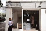 A storefront with a sign reading 'SP_CE' above the entrance, featuring a glass door and a chalkboard with text outside. A person wearing a hat and carrying a bag stands to the left, looking towards the entrance. The building is in an urban environment with nearby buildings visible in the background.