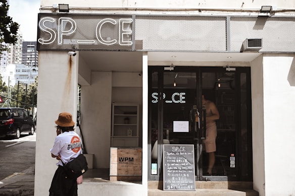 A storefront with a sign reading 'SP_CE' above the entrance, featuring a glass door and a chalkboard with text outside. A person wearing a hat and carrying a bag stands to the left, looking towards the entrance. The building is in an urban environment with nearby buildings visible in the background.