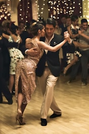 A couple is engaged in an elegant dance on a wooden floor surrounded by people. The female dancer is wearing a sparkling gold dress, and the male dancer is dressed in a dark suit with a light shirt. The atmosphere is festive, with string lights hanging in the background.