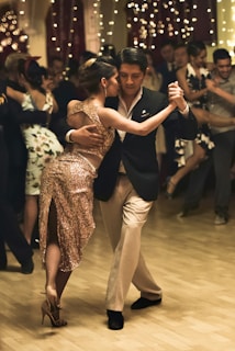 A couple is engaged in an elegant dance on a wooden floor surrounded by people. The female dancer is wearing a sparkling gold dress, and the male dancer is dressed in a dark suit with a light shirt. The atmosphere is festive, with string lights hanging in the background.
