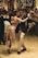 A couple is engaged in an elegant dance on a wooden floor surrounded by people. The female dancer is wearing a sparkling gold dress, and the male dancer is dressed in a dark suit with a light shirt. The atmosphere is festive, with string lights hanging in the background.