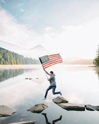 person holding us a flag standing on rock near lake during daytime