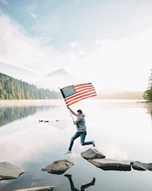 person holding us a flag standing on rock near lake during daytime