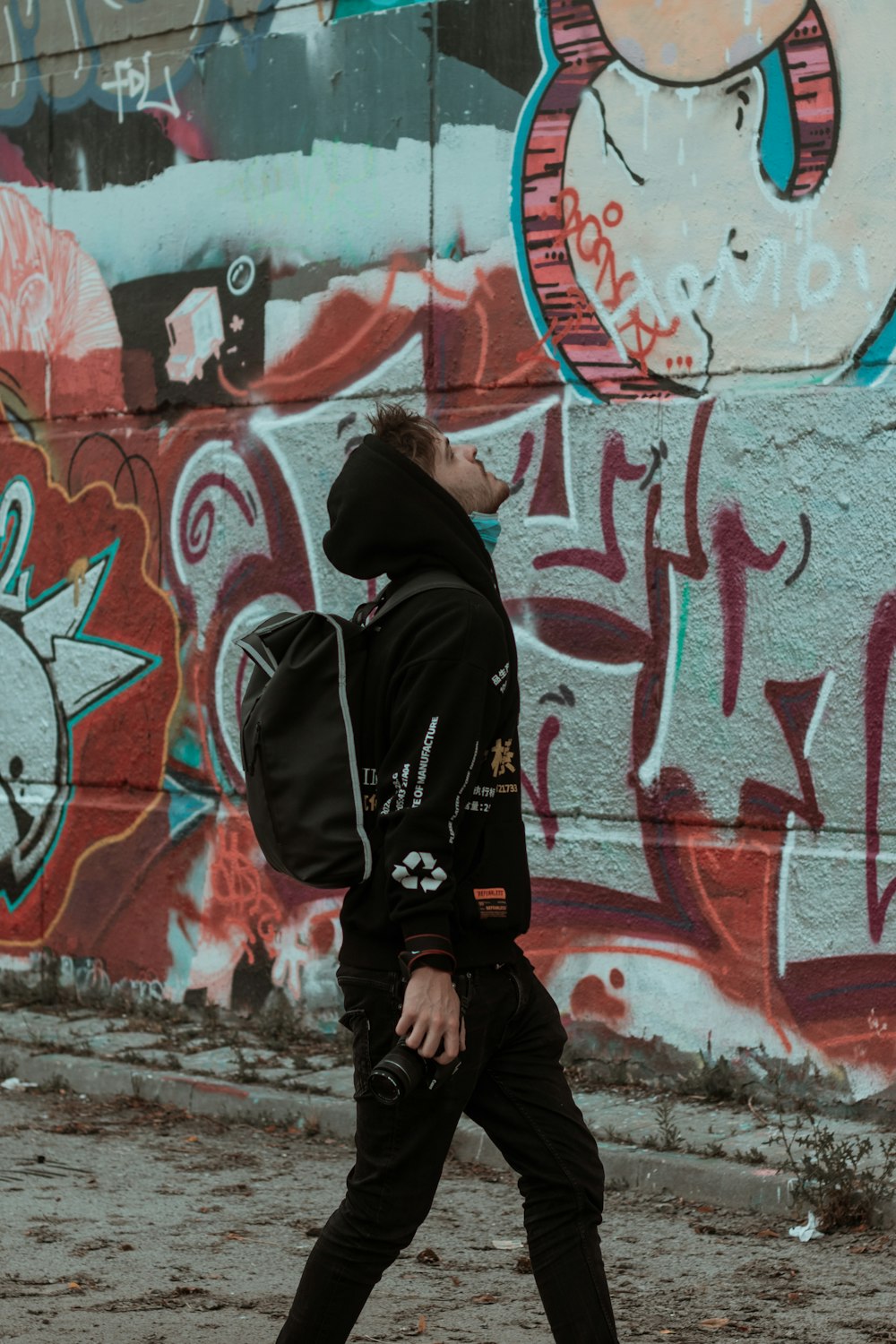 man in black hoodie and black backpack standing beside wall with graffiti