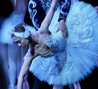 A ballet dancer is gracefully performing a move while wearing an ornate, white tutu adorned with intricate beading and patterns. The dancer's posture is elegant, with an outstretched arm and a focused expression. Another dancer's hands are gently guiding her. The background is dimly lit, highlighting the dancer's intricate costume details.