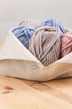 A collection of yarn balls in pastel colors is neatly placed in a light-colored fabric basket. The yarn colors include soft blue, pink, and taupe, and they have a chunky texture.
