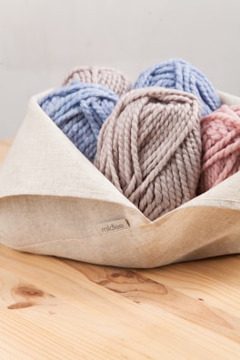 A collection of yarn balls in pastel colors is neatly placed in a light-colored fabric basket. The yarn colors include soft blue, pink, and taupe, and they have a chunky texture.