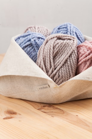 A collection of yarn balls in pastel colors is neatly placed in a light-colored fabric basket. The yarn colors include soft blue, pink, and taupe, and they have a chunky texture.