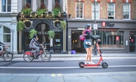 A street scene featuring a woman on an electric scooter and a person on a bicycle riding past a pub and a Vietnamese restaurant. The scooter is orange and black, and the woman is wearing a helmet, a bag over her shoulder, and casual clothing. The pub has Victorian-style architecture with plants hanging above the entrance. The bike rider is in a hoodie and casual pants, riding a branded bicycle.
