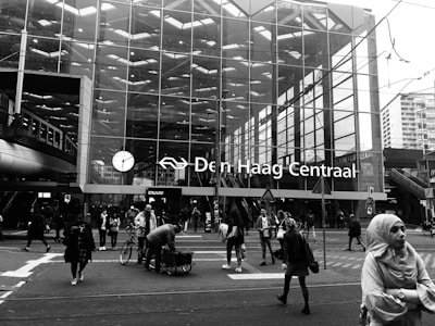 A bustling scene in front of a modern glass building with large windows, identified as 'Den Haag Centraal' station. People are walking, with some carrying bags, and one person handling a bicycle. There are visible street signs and overhead wires.