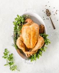 A whole roasted chicken sits in an oval white baking dish, garnished with fresh herbs such as parsley and dill. To the side, a spoon holding a mix of whole peppercorns rests on the light-colored surface.