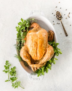 A whole roasted chicken sits in an oval white baking dish, garnished with fresh herbs such as parsley and dill. To the side, a spoon holding a mix of whole peppercorns rests on the light-colored surface.