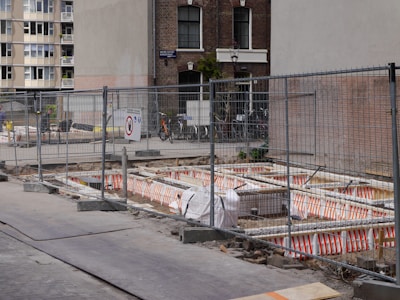 A construction site is enclosed by metal fencing, with visible orange and white safety barriers inside. There are several bicycles parked against a building in the background. A mix of residential and commercial buildings surround the site, with some windows having floral decorations.