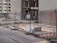A construction site is enclosed by metal fencing, with visible orange and white safety barriers inside. There are several bicycles parked against a building in the background. A mix of residential and commercial buildings surround the site, with some windows having floral decorations.