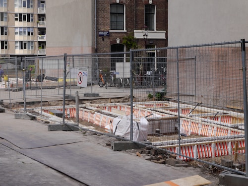 A construction site is enclosed by metal fencing, with visible orange and white safety barriers inside. There are several bicycles parked against a building in the background. A mix of residential and commercial buildings surround the site, with some windows having floral decorations.