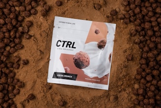 A white package labeled 'CTRL A Meal Replacement' with the flavor 'Cocoa Crunch' is placed on a bed of cocoa powder. The package design features images of cereal pieces and milk splashes. Surrounding the package are round, chocolate-colored cereal pieces.