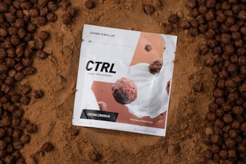 A white package labeled 'CTRL A Meal Replacement' with the flavor 'Cocoa Crunch' is placed on a bed of cocoa powder. The package design features images of cereal pieces and milk splashes. Surrounding the package are round, chocolate-colored cereal pieces.