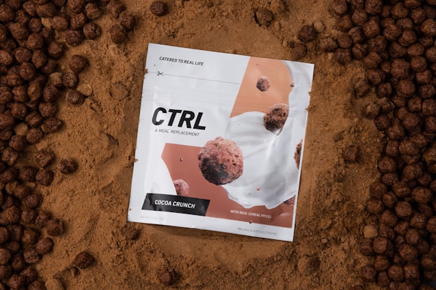 A white package labeled 'CTRL A Meal Replacement' with the flavor 'Cocoa Crunch' is placed on a bed of cocoa powder. The package design features images of cereal pieces and milk splashes. Surrounding the package are round, chocolate-colored cereal pieces.