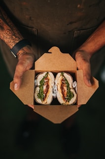 A person is holding an open cardboard box containing two neatly arranged sandwiches. The sandwiches have visible ingredients like greens and meat, and are wrapped with some paper underneath. The person has tattoos on one arm and is wearing a dark-colored apron.